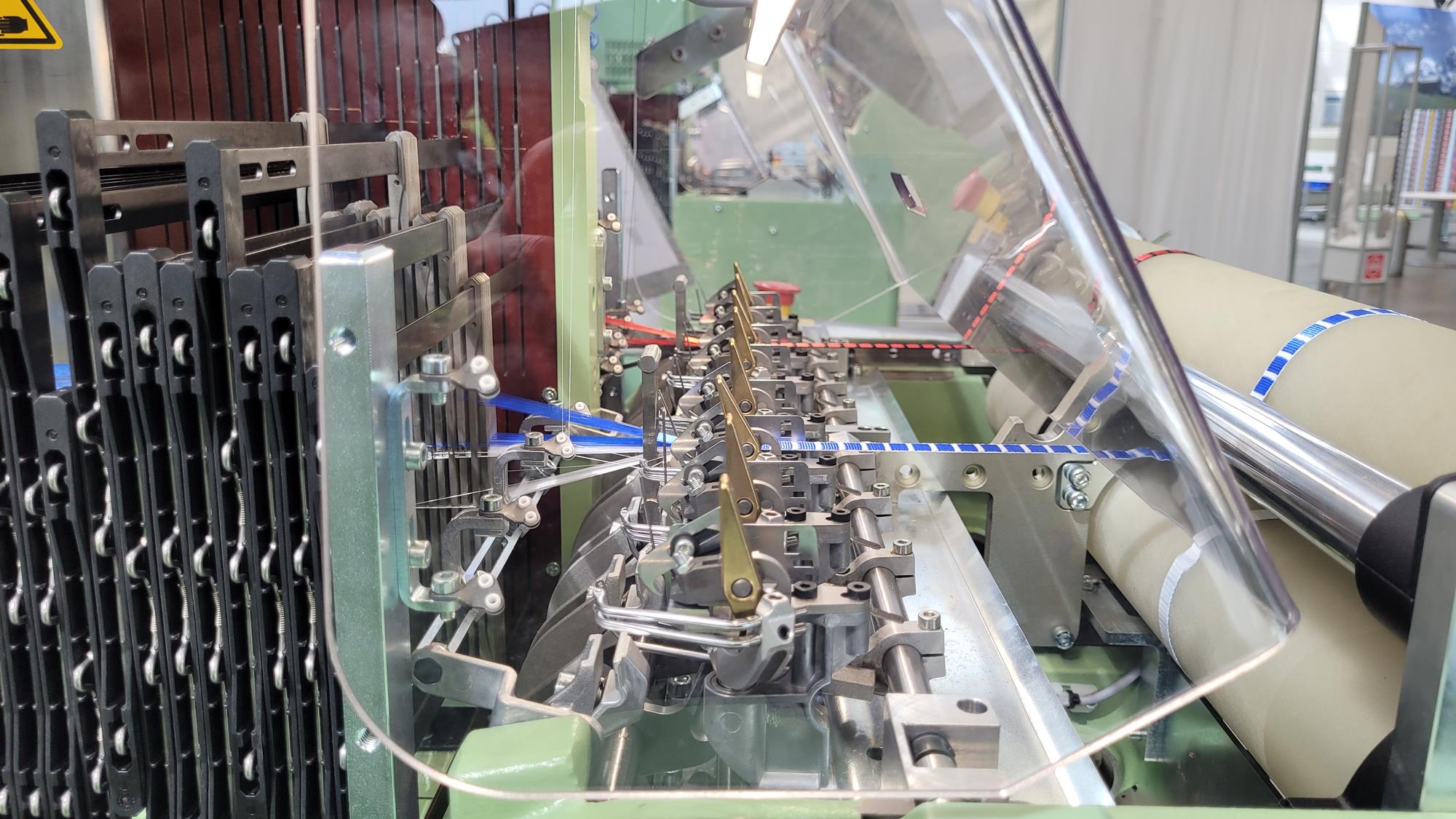 Detailansicht einer textilproduzierenden Maschine der Firma Jakob Müller aus Frick, mit sichtbaren Spulen, Fäden und mechanischen Teilen.