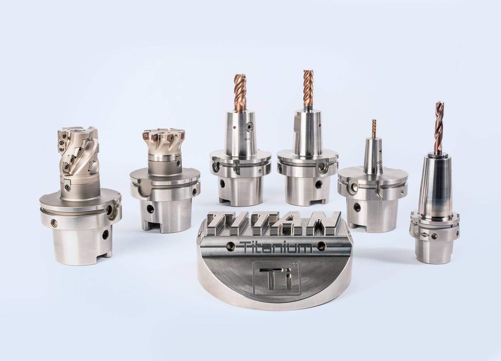 Eine Sammlung von präzisionsgefertigten Metallwerkzeugen und -komponenten für die maschinelle Bearbeitung auf einem weißen Hintergrund. Im Vordergrund liegt eine größere runde metallene Vorrichtung mit eingraviertem Wort "Titanium".